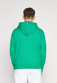 Grüne Kapuzenjacke aus strukturiertem Stoff, mit einer Schnürung an der Kapuze und einem kleinen, dreifarbigen Akzent am Hals. Glatte Oberfläche, normale Passform.