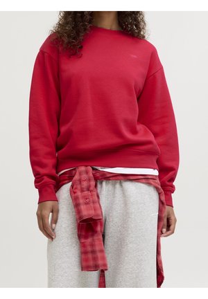 Personne portant un sweat-shirt rouge, un pantalon de jogging gris et une chemise à carreaux rouge nouée autour de la taille, debout devant un fond clair uniforme.