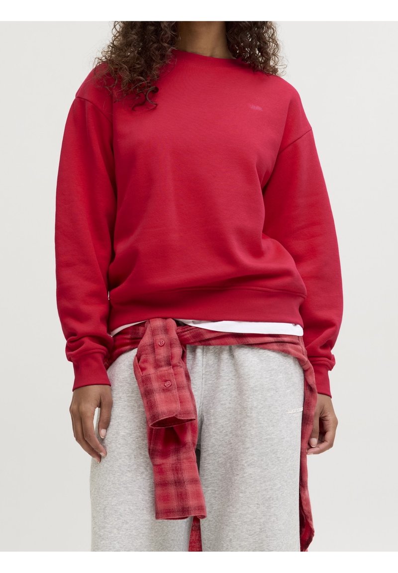 Personne portant un sweat-shirt rouge, un pantalon de jogging gris et une chemise à carreaux rouge nouée autour de la taille, debout devant un fond clair uniforme.
