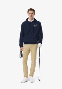 Felpa blu marina con scritte bianche, pantaloni beige e scarpe da golf bianche. Tiene in mano una borsa a rete con palline da golf e un bastone da golf.