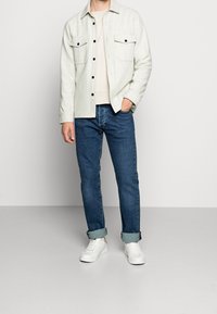 Ljusgrå, skjorta med knappar och två bröstfickor, tillsammans med mörkblå jeans. Vita sneakers fulländar outfiten.