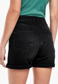 Černé džíny šortky s ohrnutým lemem, které mají dvě zadní kapsy. Materiál je hladký s mírným leskem a těsný střih.