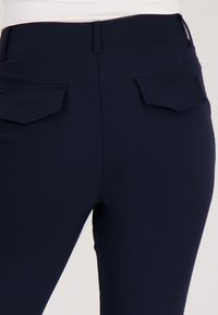 Donkerblauwe nauwsluitende broek met een gladde textuur, voorzien van twee hoekige achterzakken en een ritsdetail bij de taille.
