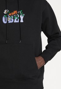 Svart hoodie med färgglad broderi av blommor och en sko, med en känguruficka och dragsnören på huvan.