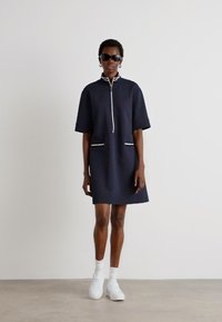 Navy short-sleeve dress with two front pockets, white trim, and a zip neck. Worn with white sneakers and socks, against a plain background.