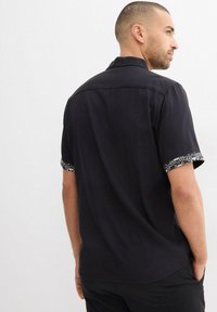 Maglietta nera a maniche corte con una vestibilità rilassata. Presenta polsini a fantasia e una texture liscia. Colletto a bottoni e dettagli sulla parte posteriore.