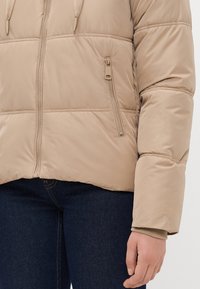 Beez puffer jakker, mille sileda tekstuuriga, esiküljel tõmblukk ja küljetasku. Kaunistatud elastsete varrukatega ja lühikese pikkusega.