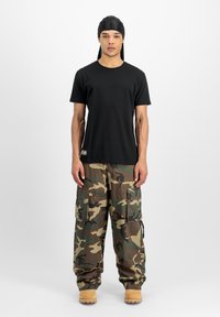 T-shirt en coton noir associé à un pantalon cargo camouflage vert et marron. Le pantalon possède de multiples poches et une coupe ample. Des bottes marron complètent la tenue.