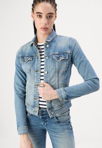 Light blue denim jacket featuring a fitted design, button closures, chest pockets, and a slightly faded texture. Striped top beneath.