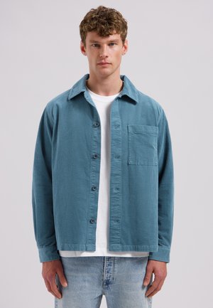 Ung mand iført en blå fløjlsbluse over en hvid T-shirt og lyseblå denimjeans, stående foran en grå baggrund.