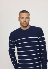 Pull en marine avec des rayures blanches horizontales, col rond, poignets et ourlet côtelés, comprenant une petite étiquette en cuir marron sur le côté.