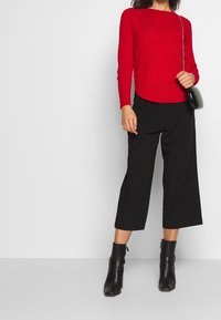 Röd stickad tröja med ett texturerat mönster, matchad med svarta vida byxor. Modellen har på sig svarta ankelstövlar och håller en liten svart väska.