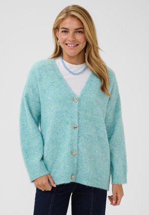 Femme souriante aux longs cheveux blonds porte un cardigan duveteux bleu clair avec des boutons argentés, une chemise blanche, un collier bleu et un jean foncé.