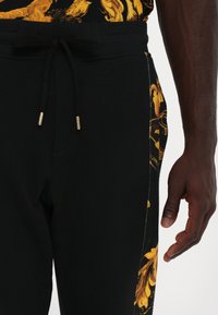 Black joggers with a drawstring waist and golden floral pattern on the side. Features a smooth texture and casual fit.