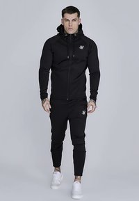 Sudadera negra con cremallera y diseño ajustado, con un logo en el pecho y capucha con cordones. Combinada con pantalones joggers negros a juego y zapatillas blancas.