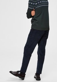 Mörkblå tapered-byxor gjorda av mjukt tyg, kombinerade med en mörkgrön tröja med vita geometriska mönster och ribbade muddar. Svarta skor.