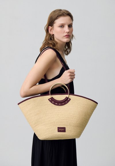 Sac de plage tressé en paille avec une forme large, mettant en avant des accents en cuir bordeaux et une poignée en métal circulaire. Surface texturée avec un branding subtil.