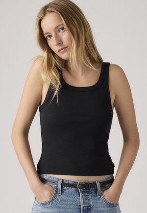 Νεαρή γυναίκα με μακριά ξανθά μαλλιά, ντυμένη με μαύρο αμάνικο μπλουζάκι και μπλε τζιν με μαύρη ζώνη, με τα χέρια στις τσέπες, ουδέτερο φόντο.