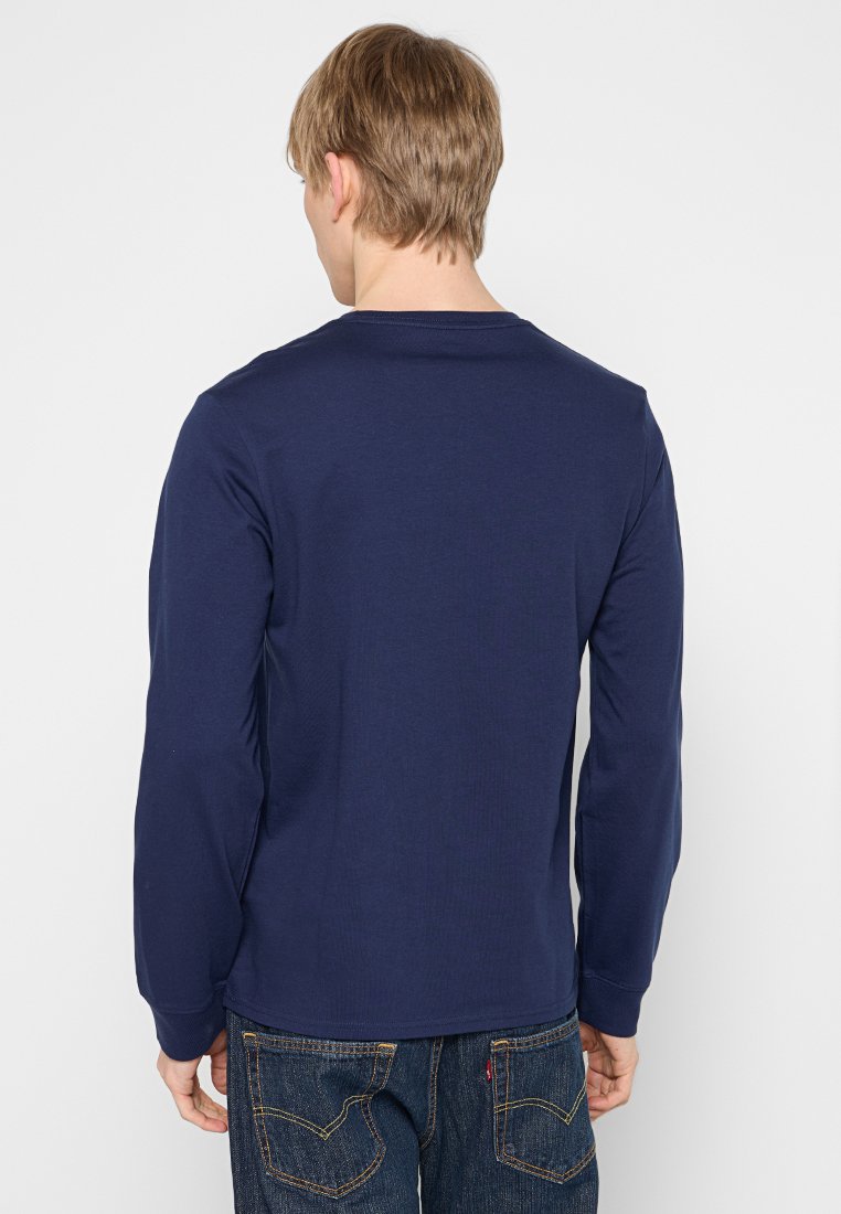 Hombre con cabello rubio, vistiendo una camisa de manga larga azul marino y jeans azules, de pie, de espaldas, frente a un fondo blanco liso.