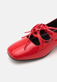 Red patent leather ballet shoe with a rounded toe, featuring a cutout design and matching bow laces across the top. Black insole.