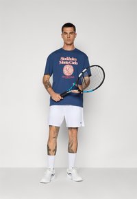 Marineblå t-shirt med rød grafisk print, hvide shorts, hvide sneakers og blå tennisracket. Mandlig figur med tatoveringer står på en ensfarvet baggrund.
