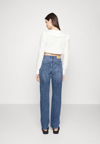 Camisa branca cropped de manga longa com colarinho franzido, emparelhada com jeans de denim azul de cintura alta e perna reta. Sapatos pretos visíveis.