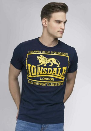 Marineblaues Baumwoll-T-Shirt mit einem abgenutzten gelben Grafikdruck eines Löwen und dem Text "Lonsdale London" auf der Vorderseite. Reguläre Passform, kurze Ärmel.