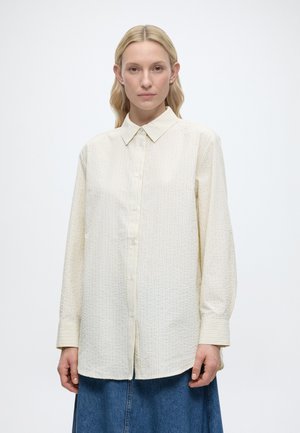 Ξανθιά γυναίκα που φορά μπλούζα με μακριά μανίκια, κρεμ με ρίγες και μπλε τζιν φούστα, σε απλό λευκό φόντο.