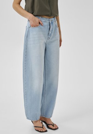 Persoon draagt lichtblauwe jeans met wijde pijpen, olijfgroene cropped top en zwarte sandalen met hak, staat met één hand in de zak tegen een witte achtergrond.