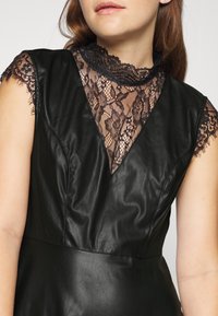 Femme portant une robe sans manches en similicuir noir avec un col haut en dentelle noire transparente et des manches courtes.