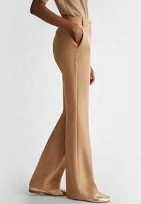 Beige hoge taillebroek met uitlopende pijpen en zijzakken; gladde textuur; gecombineerd met glanzende gouden flats.