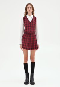 Gilet a quadri rossi e neri abbinato a una gonna plissettata coordinata. Camicia bianca a maniche lunghe sotto il gilet. Calze nere al ginocchio e stivali robusti.