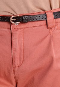 Rust-colored cotton pants with a smooth texture, featuring front pockets, held up by a black perforated leather belt with a silver buckle.