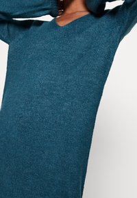 Maglione teal a scollatura a V realizzato in morbido tessuto lavorato a maglia. Presenta maniche lunghe e una vestibilità comoda con una leggera texture su tutta la superficie.