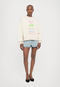 Krémová mikina s farebným potlačeným textom, spárovaná so svetlomodrými rifľovými šortkami. Model má na sebe čierne baleríny, na neutrálne pozadie.