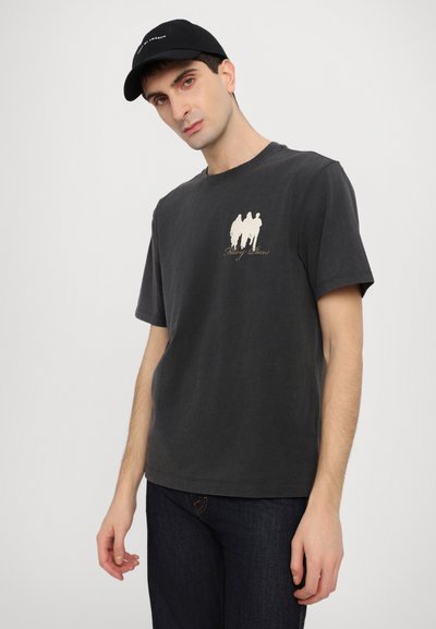 Black cotton t-shirt featuring a cream graphic of three people and text, short sleeves, round neck, paired with a black cap.