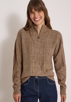 Femme souriante, portant un pull marron en maille torsadée zippé et un jean bleu foncé, debout devant un fond beige uni.