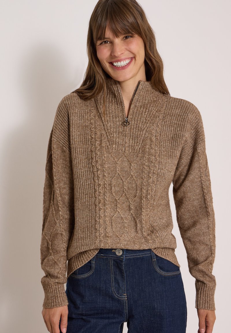 Femme souriante, portant un pull marron en maille torsadée zippé et un jean bleu foncé, debout devant un fond beige uni.
