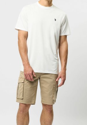 Homme portant un t-shirt blanc uni à manches courtes avec un petit logo noir et un short cargo beige avec de grandes poches latérales.