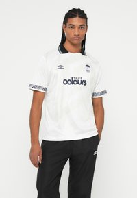 Jauns vīrietis, tērpies baltā Rainy City FC futbola kreklā ar melnu apkakli un apmalēm, apvienots ar melniem Umbro sporta biksēm, stāv uz balta fona.
