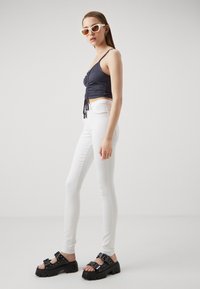 Merine ribbed lühike topp koos kogutud detailidega, kombineeritud valgete kitsaste teksadega ja mustade jämedate sandaalidega, millel on nõelad ja pandlad.