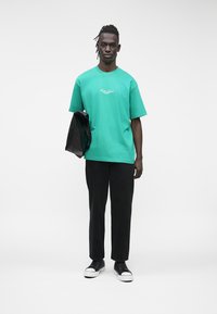 Zaļš kokvilnas T-krekls ar īsām piedurknēm un logotipa detaļu, kombinēts ar melnām biksēm un melnām augstām sporta kurpēm. Modelis tur melnu jaku.