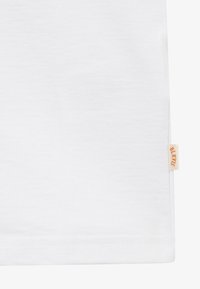 Tissu blanc avec une petite étiquette beige sur le bord droit, avec un texte orange indiquant "BLKVIS" près de l'ourlet inférieur.