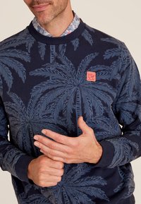 Sweat-shirt bleu foncé avec un motif de feuille de palmier en bleu clair. Il présente un col et des poignets côtes, avec une petite étiquette de marque rouge sur la poitrine.