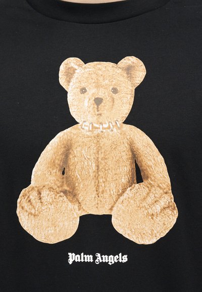 Brauner Teddybär mit einer Halskette, der vor einem schwarzen Hintergrund sitzt, darunter der Schriftzug "Palm Angels" in weißer gotischer Schrift.