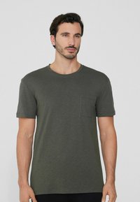 Oliwkowy t-shirt z krótkim rękawem wykonany z miękkiego materiału, z okrągłym dekoltem i kieszonką na lewej piersi. Gładka tekstura, luźny krój.