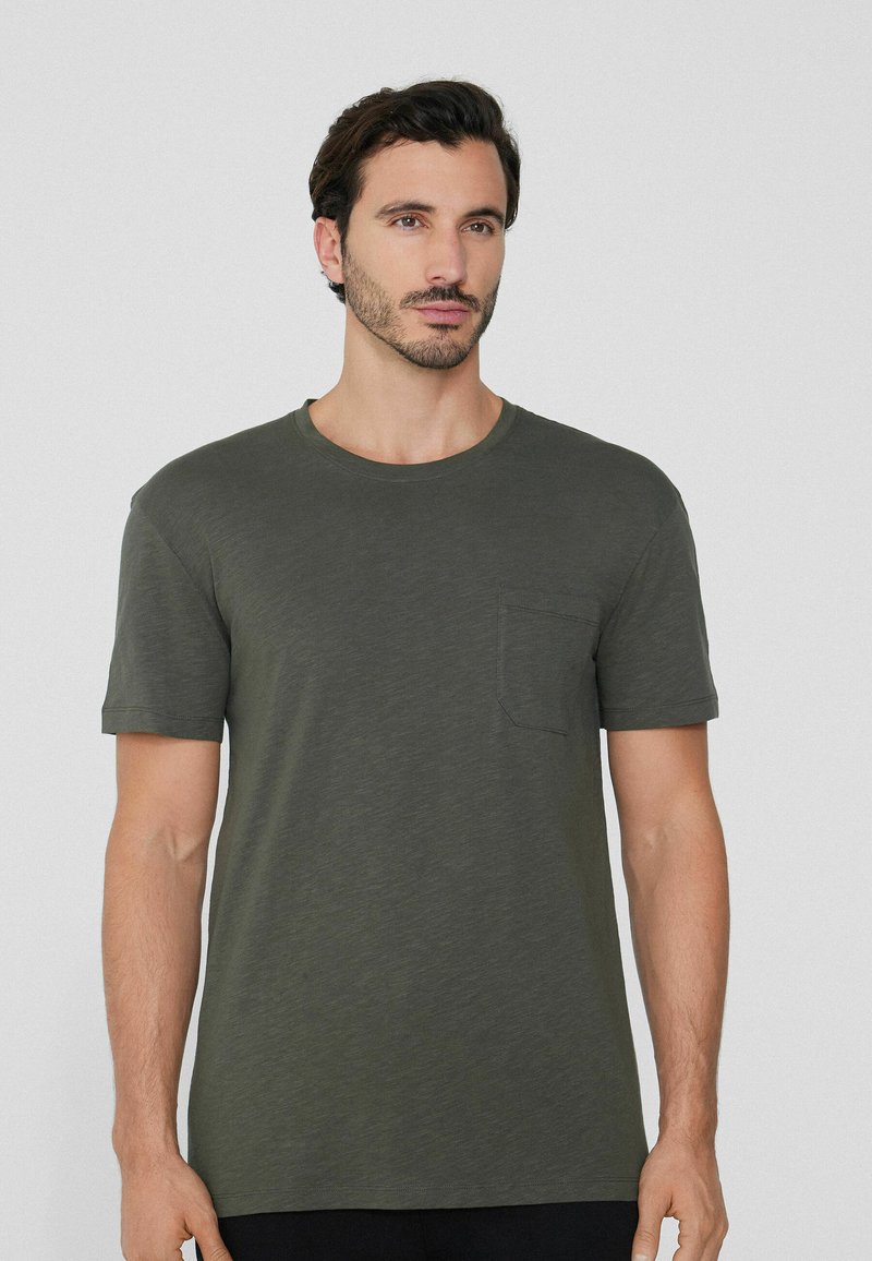 Oliwkowy t-shirt z krótkim rękawem wykonany z miękkiego materiału, z okrągłym dekoltem i kieszonką na lewej piersi. Gładka tekstura, luźny krój.