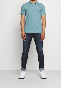 Polo turquoise avec col, manches courtes et logo, associé à un jean denim bleu foncé et des baskets blanches.