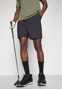 Calças de golfe pretas com uma textura suave, combinadas com uma camisa verde, meias pretas e sapatos de golfe pretos. Taco de golfe segurado numa mão.