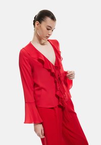 Blouse rouge avec encolure en V à volants et longues manches en cloche. Fabriquée dans un tissu léger avec un détail noué à l'avant. Texture douce.